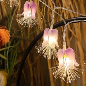 Puede incluir: Una guirnalda de luces decorativas con cubiertas de flores translúcidas en forma de campana en tonos blanco y morado. Cada flor está acentuada con un grupo de filamentos de fibra óptica finos y radiantes que brillan al iluminarse.