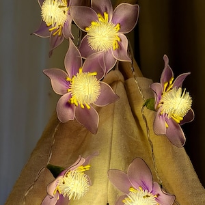 May include: A string of decorative flower lights. Each flower has purple petals, a yellow center, and yellow accents. The lights are strung on a thin wire and are arranged in front of a tan fabric.