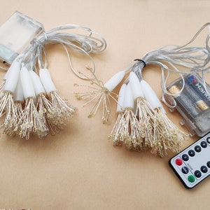 K&ouml;nnte beinhalten: Ein Satz wei&szlig;er Lichterketten mit einer Fernbedienung. Die Lichter sind batteriebetrieben und haben einen warmwei&szlig;en Schein. Die Fernbedienung hat Kn&ouml;pfe f&uuml;r Ein/Aus, Timer und verschiedene Lichtmodi.