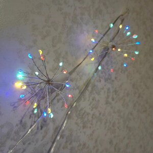 K&ouml;nnte beinhalten: Zwei Lichterketten mit mehrfarbigen Gl&uuml;hbirnen, die wie ein Feuerwerk geformt sind, h&auml;ngen von einer Decke.