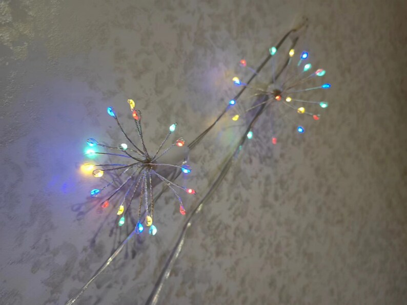 K&ouml;nnte beinhalten: Zwei Lichterketten mit mehrfarbigen Gl&uuml;hbirnen, die wie ein Feuerwerk geformt sind, h&auml;ngen von einer Decke.