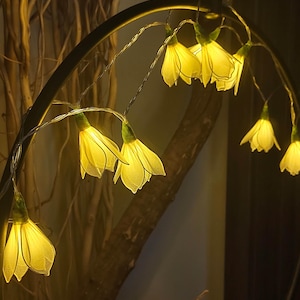 May include: A string of decorative fairy lights shaped like yellow flowers. The lights are strung on a thin, silver wire and are illuminated, creating a warm, inviting glow. The background includes a dark, curved metal structure and a tree branch.