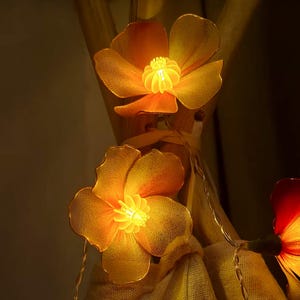 May include: Close-up of a string of decorative lights featuring glowing orange flower-shaped bulbs. The flower petals have a mesh-like texture, and the centers emit a bright, warm light. The string lights are ideal for adding a touch of ambiance.