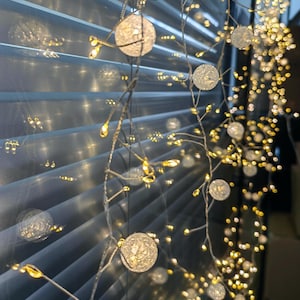May include: String lights with small, round, textured, white orbs and tiny yellow lights. The lights are strung on thin, silver wires. The background shows a window with closed blinds, creating a soft, diffused light effect. The lights are likely for decorative purposes.