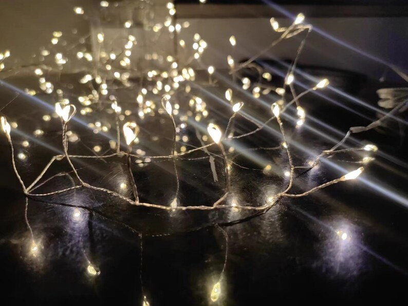 Pu&ograve; includere: Una ghirlanda di luci bianche calde con un filo di rame, disposte in un motivo fantasioso. Le luci sono infilate su una superficie scura, creando un'atmosfera calda e invitante.