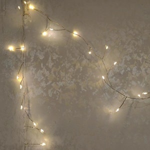 Pu&ograve; includere: Una ghirlanda di luci bianche calde avvolte attorno a un muro con una texture. Le luci sono accese e creano un bagliore morbido.