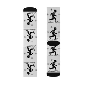 Op de afbeelding: Een paar witte sokken met zwarte illustraties van skiërs en sneeuwvlokken. De sokken hebben een zwarte boord en teen. De skiër illustraties worden herhaald over de lengte van de sokken.