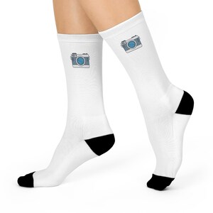 Op de afbeelding: Een paar witte sokken met een zwarte hiel en teen. Elke sok heeft een klein, cartoonachtig cameraontwerp in grijs en blauw.