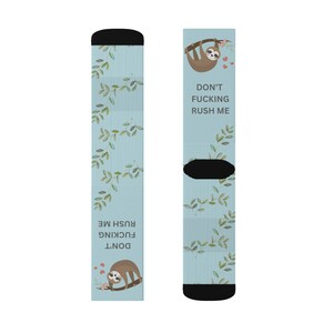 Könnte beinhalten: Hellblaue Socken mit einem Faultiermotiv und dem Text "DON'T FUCKING RUSH ME". Die Socken zeigen ein Faultier, das an einem Ast mit Blättern und kleinen roten Herzen hängt. Die Oberseite der Socken ist schwarz.