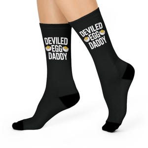 Peut inclure: Chaussettes noires avec l'inscription "DEVILED EGG DADDY" en blanc, accompagnée d'un dessin d'œuf farci. Les chaussettes sont portées aux pieds.