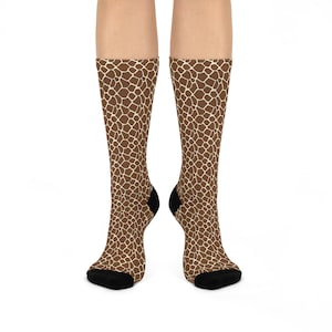May include: A pair of knee-high socks featuring a giraffe print pattern in shades of brown and cream. The socks have black heels and toes, and are displayed against a white background.