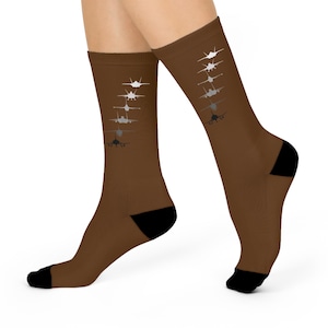 Puede incluir: Calcetines marrones con talones y punteras negros. Los calcetines presentan un patrón repetido de siluetas de aviones en blanco y gris, creando un diseño dinámico. Los calcetines se llevan en las piernas de una persona.