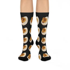 Peut inclure: Chaussettes noires montantes avec un motif répété d'œufs farcis. Chaque œuf est représenté avec un blanc d'œuf blanc et un jaune d'œuf jaune-orangé. Les chaussettes sont portées sur les jambes d'une personne.
