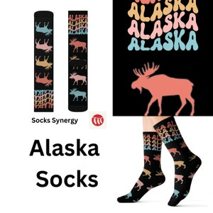 May include: Black socks with colorful moose silhouettes and the word "ALASKA" repeated in a retro font. The socks are displayed on a white background, with the text "Alaska Socks" below. The brand name "Socks Synergy" is also visible.