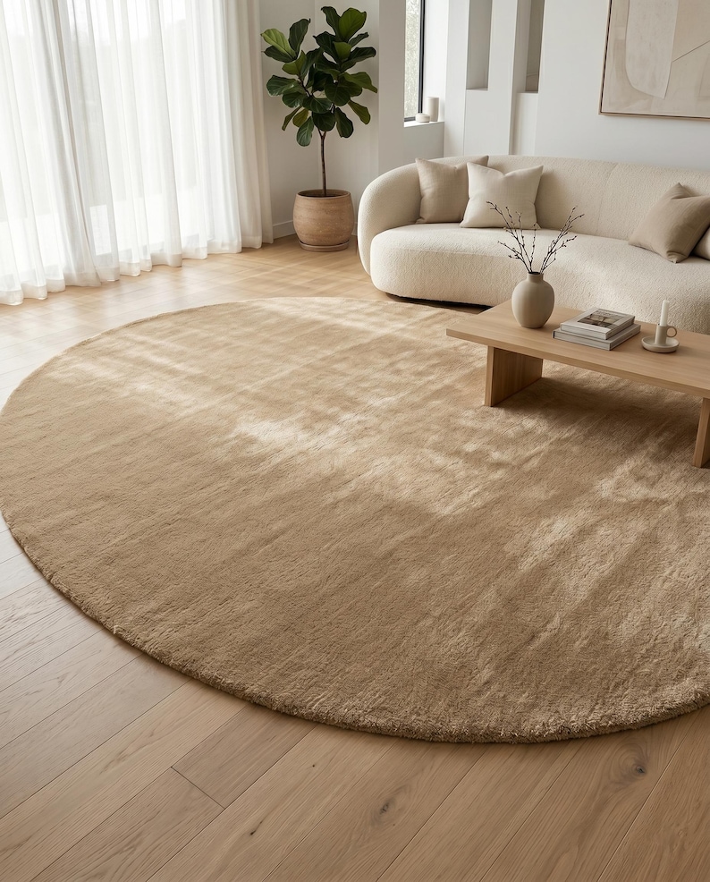 Modern beige hand tufted rug displayed in a designer-inspired living room featuring oversized sectional seating, natural wood coffee tables, soft textiles, indoor plants, and abundant natural lighting. The plush wool surface introduces subtle texture and visual warmth while the neutral sand beige palette complements Scandinavian, Japandi, contemporary, organic modern, and luxury interior styles with effortless elegance.