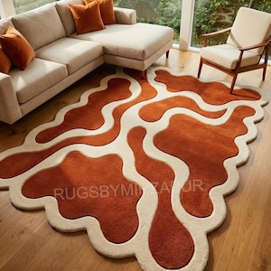 Hand-tufted wool rug in an organic amoeba shape featuring flowing rust orange fields divided by soft ivory wave channels. The rug has a deep high-low carved texture creating a strong 3D effect. Styled in a modern living room with beige sectional sofa, wooden flooring, warm cushions, and large windows overlooking greenery.