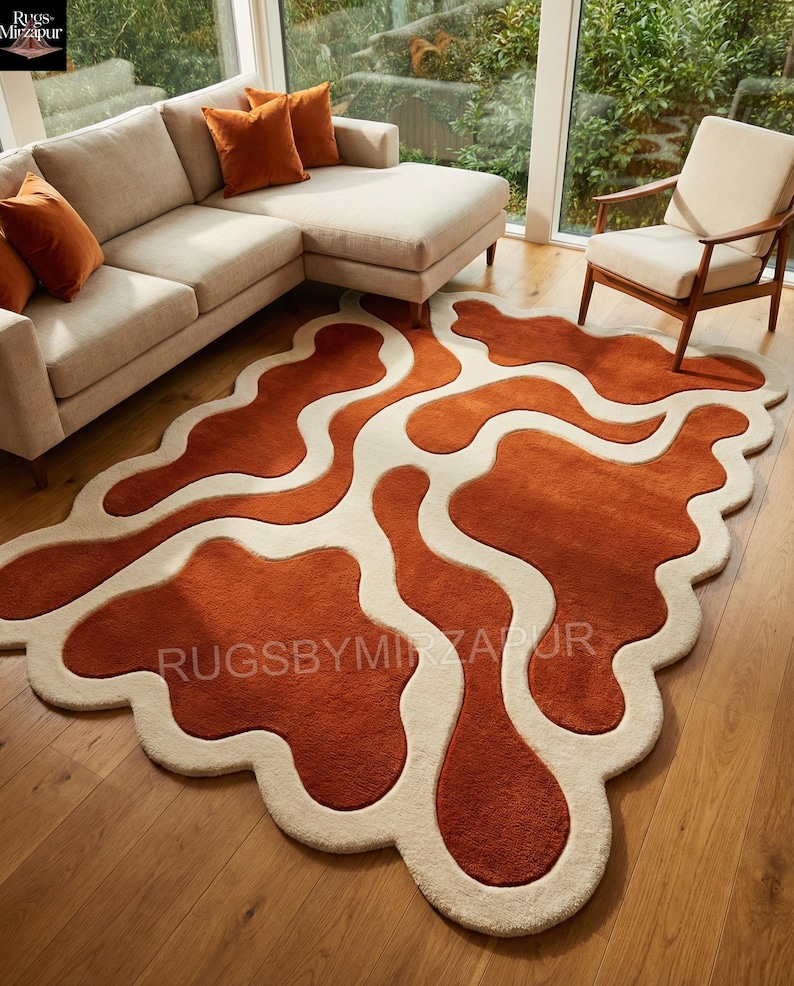 Hand-tufted wool rug in an organic amoeba shape featuring flowing rust orange fields divided by soft ivory wave channels. The rug has a deep high-low carved texture creating a strong 3D effect. Styled in a modern living room with beige sectional sofa, wooden flooring, warm cushions, and large windows overlooking greenery.