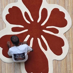 May include: A large, flower-shaped rug with a rust-colored center and off-white petals. The rug is on a light brown wooden floor. A person wearing a white t-shirt is sitting on the floor near the rug.