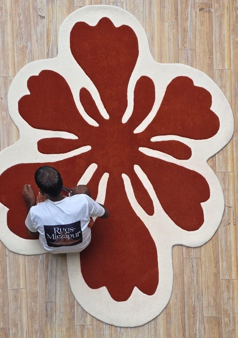 May include: A large, flower-shaped rug with a rust-colored center and off-white petals. The rug is on a light brown wooden floor. A person wearing a white t-shirt is sitting on the floor near the rug.