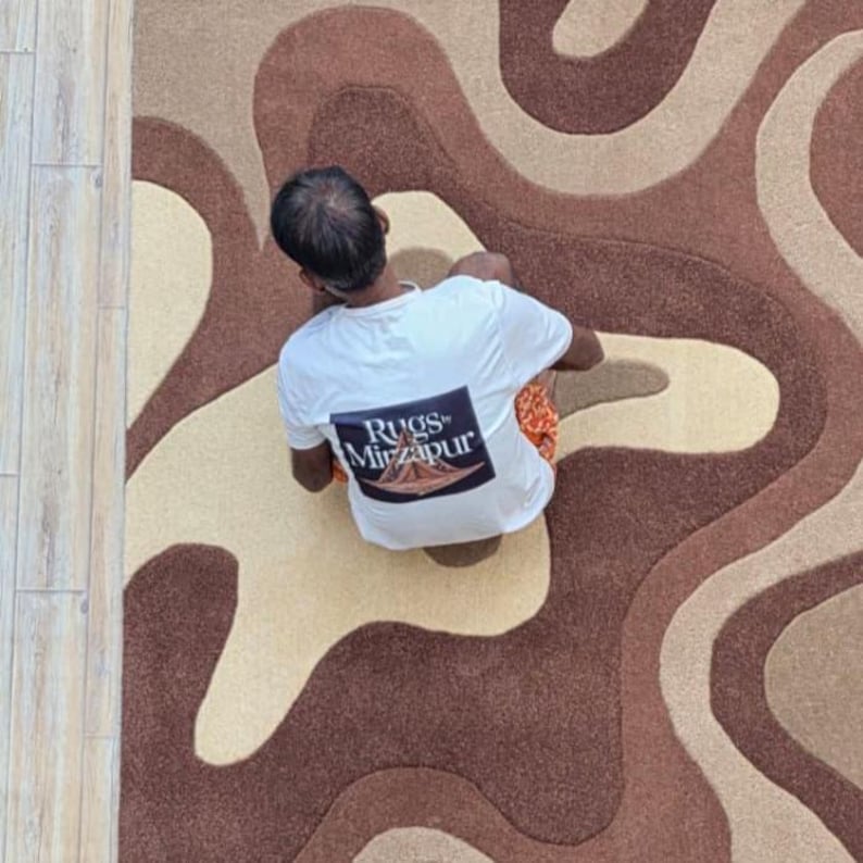 Op de afbeelding: Luchtfoto van een tapijt met een bruin, beige en tan golvend patroon. Een persoon die een wit t-shirt draagt met de tekst "Rugs Mirzapur" zit op het tapijt. Het tapijt heeft een abstract ontwerp.