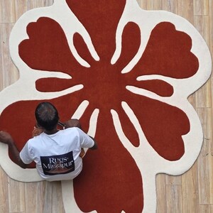 May include: A large, flower-shaped rug with a rust-colored center and a cream-colored border. The rug is on a light brown wooden floor. The text "Rugs Mirzapur" is visible on a person's shirt.