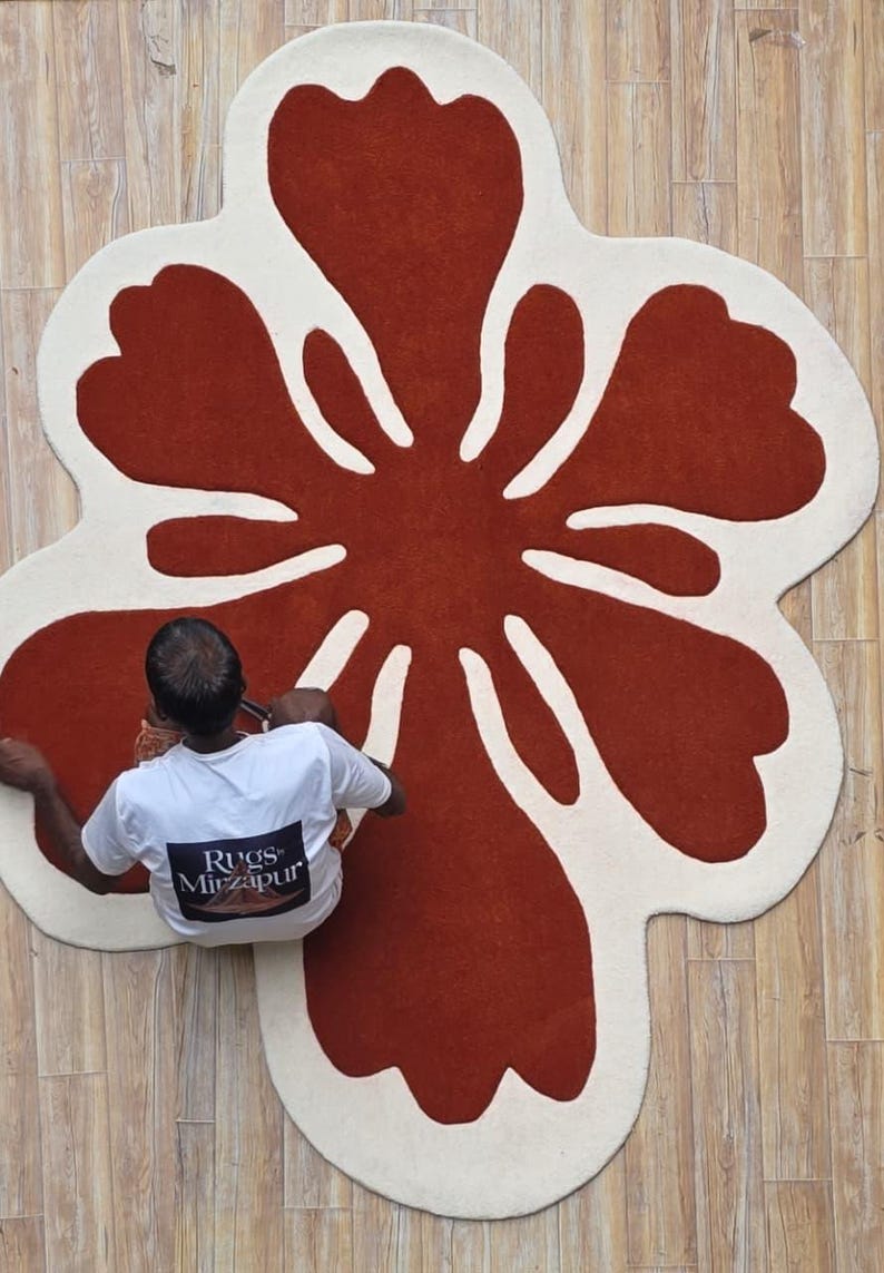 May include: A large, flower-shaped rug with a rust-colored center and a cream-colored border. The rug is on a light brown wooden floor. The text "Rugs Mirzapur" is visible on a person's shirt.