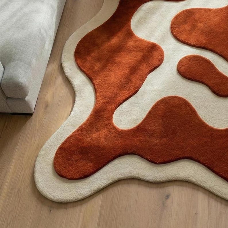 Modern hand-tufted wool area rug in an irregular organic shape, designed with flowing terracotta abstract forms layered over an ivory background. Dense plush wool pile with hand-carved contours creates a three-dimensional effect. Perfect for living rooms, bedrooms, or studios, this rug complements minimalist, mid-century, and contemporary home décor styles.