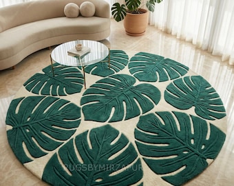 May include: A round rug featuring a pattern of large, dark green monstera leaves against a cream-colored background. The rug is centered in a room with a curved beige sofa, a glass-topped coffee table, and a potted plant. The text "RUGSBYMIRZAPUR" is visible on the rug.