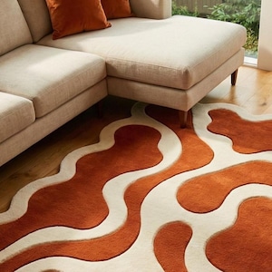 Luxury hand-tufted wool rug featuring flowing wave patterns in rust orange and ivory. The irregular amoeba shape and deep carved pile create a tactile, sculptural look. Shown in a contemporary interior with a corner sofa, accent chair, and garden views through large windows.