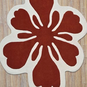 May include: A large, flower-shaped rug with a bold design. The rug features a deep red center with white petal outlines. The rug is placed on a light-colored wooden floor, creating a striking contrast.