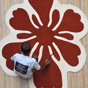 May include: A large, flower-shaped rug with a rust-colored center and off-white petals. The rug is on a light-colored wooden floor. A person is working on the rug with a knife. The person is wearing a white t-shirt with the text "Rugs Mirzapur".