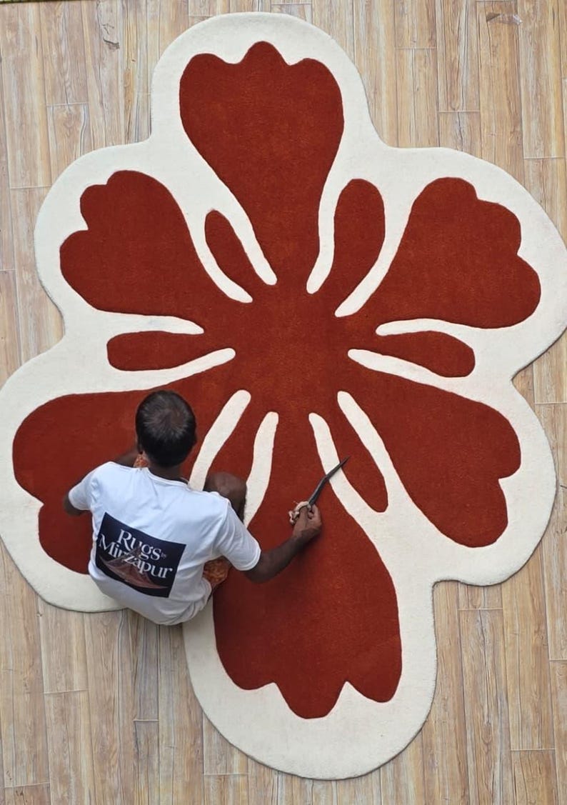 May include: A large, flower-shaped rug with a rust-colored center and off-white petals. The rug is on a light-colored wooden floor. A person is working on the rug with a knife. The person is wearing a white t-shirt with the text "Rugs Mirzapur".