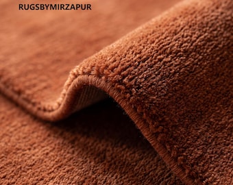 May include: A solid rust-colored rug with a dense, short-pile texture and a folded edge. The text RUGSBYMIRZAPUR is printed in black on the upper left side of the rug. The surface features a uniform, soft, and fuzzy appearance throughout the piece.