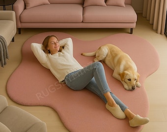 May include: A large, irregularly shaped, blush pink rug dominates the scene. A woman in a white sweater and jeans relaxes on the rug, with a golden Labrador dog nearby. A pink sofa and pillows are in the background. The rug has a soft, plush texture.