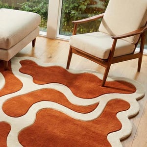 Designer organic wool rug with abstract river-like wave design in rust and ivory shades. Positioned in a modern living room with light furniture, wooden flooring, and soft daylight. The plush high-low pile adds dimension and artistic character.