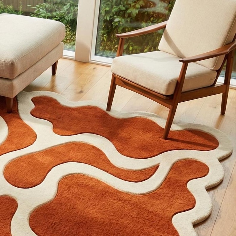 Designer organic wool rug with abstract river-like wave design in rust and ivory shades. Positioned in a modern living room with light furniture, wooden flooring, and soft daylight. The plush high-low pile adds dimension and artistic character.