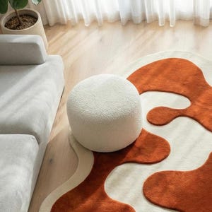 Luxury handmade wool rug featuring bold terracotta abstract motifs carved in high-low relief against a warm ivory base. Crafted using traditional tufting techniques, this sculptural rug offers a soft underfoot feel and artistic texture. Shown in a bright modern interior with neutral furniture, highlighting the rug as a statement centerpiece.