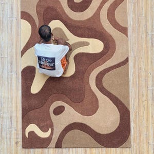 Op de afbeelding: Een groot, rechthoekig tapijt met een abstract ontwerp in bruin- en beigetinten. Het tapijt heeft een golvend patroon, waardoor een gevoel van beweging ontstaat. De tekst "Rugs-Mirzapur" is zichtbaar op een wit T-shirt.