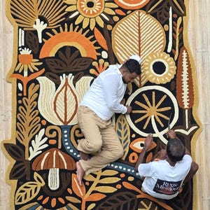 Hand-tufted wool wall rug featuring folk botanical artwork with flowers, leaves, mushrooms, and sun motifs in warm earthy colors, displayed above a modern sofa.