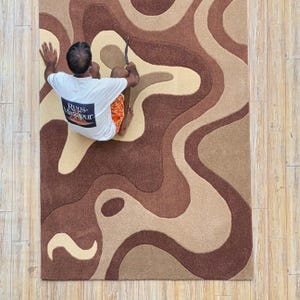 Op de afbeelding: Een bruin en beige vloerkleed met een golvend, abstract ontwerp. Het vloerkleed heeft verschillende tinten bruin en beige, waardoor een textuur ontstaat. De tekst "Rugs Mirzapur" is zichtbaar op een wit t-shirt. Het vloerkleed ligt op een houten vloer.