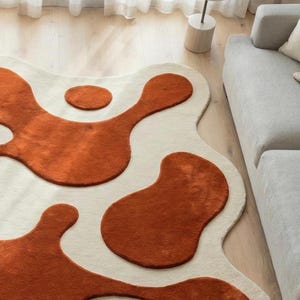 Contemporary abstract wool area rug with flowing terracotta shapes on an ivory background, hand-tufted and carved for a high-low 3D effect. Styled in a minimalist living room with soft furnishings and natural light, this designer rug adds visual movement, cozy comfort, and artistic sophistication to modern interiors.