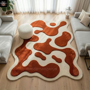 Hand tufted abstract wool rug with organic freeform shape, featuring raised terracotta and rust-colored motifs on a soft ivory base. The rug showcases a high-low carved pile creating sculptural depth and texture. Styled in a modern living room with neutral sofas, wooden flooring, and natural daylight, this artisan rug adds warmth, movement, and contemporary artistic appeal to the interior.