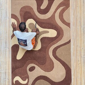 Op de afbeelding: Een rechthoekig tapijt met een vloeiend, abstract ontwerp in bruin- en beigetinten. Het tapijt heeft een golvend patroon, wat een moderne en artistieke uitstraling cre&euml;ert. Het tapijt is gemaakt van een zacht materiaal, perfect voor een woon- of slaapkamer.