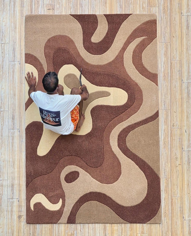 Op de afbeelding: Een rechthoekig tapijt met een vloeiend, abstract ontwerp in bruin- en beigetinten. Het tapijt heeft een golvend patroon, wat een moderne en artistieke uitstraling cre&euml;ert. Het tapijt is gemaakt van een zacht materiaal, perfect voor een woon- of slaapkamer.