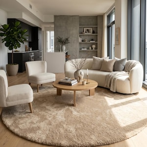 Large beige round hand tufted wool rug displayed in an open-concept living room with curved sectional seating, light wood flooring, indoor greenery, and contemporary architecture. The luxurious plush pile provides exceptional comfort while the elegant neutral color palette blends beautifully with Scandinavian, Japandi, organic modern, coastal, and contemporary interiors. This handmade area rug creates a refined foundation that enhances every designer-inspired living space.