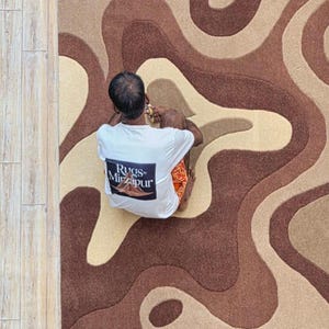 Op de afbeelding: Een bovenaanzicht van een tapijt met een bruin, beige en tan golvend patroon. Een persoon met een wit t-shirt met de tekst "Rugs of Mirzapur" zit op het tapijt.