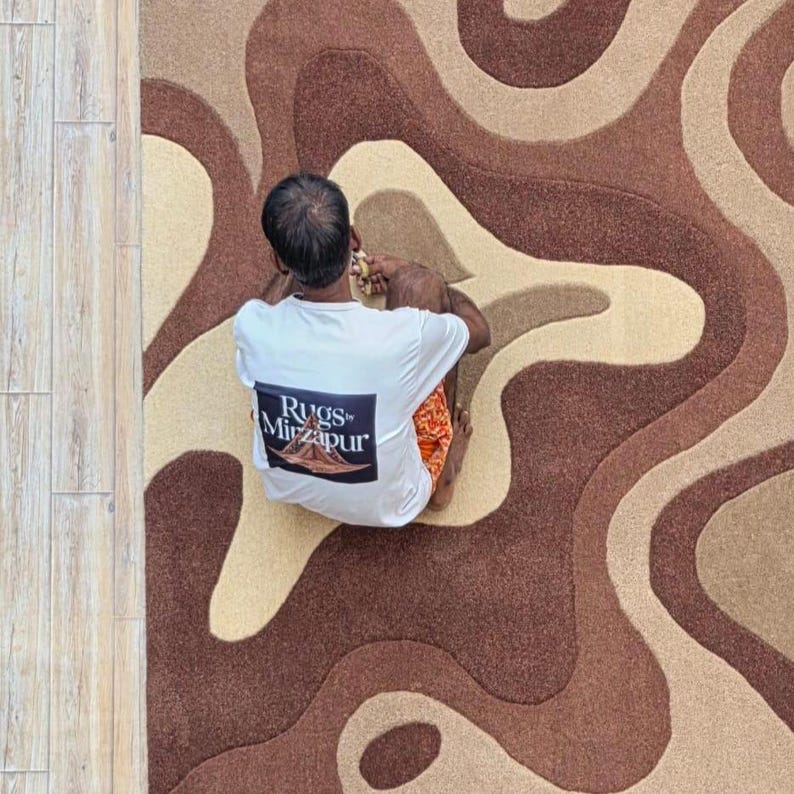 Op de afbeelding: Een bovenaanzicht van een tapijt met een bruin, beige en tan golvend patroon. Een persoon met een wit t-shirt met de tekst "Rugs of Mirzapur" zit op het tapijt.