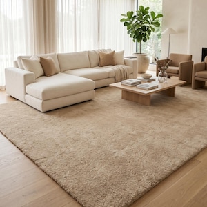 Handmade beige area rug styled in a sophisticated contemporary living room with cream upholstered seating, walnut furniture, soft neutral textiles, oversized windows, and indoor plants. The plush wool construction provides luxurious comfort while the elegant sand beige tone establishes a calm and welcoming atmosphere. Perfect for Scandinavian, Japandi, minimalist, organic modern, and luxury interiors, this hand tufted rug combines timeless craftsmanship with modern elegance.