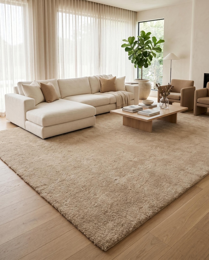 Handmade beige area rug styled in a sophisticated contemporary living room with cream upholstered seating, walnut furniture, soft neutral textiles, oversized windows, and indoor plants. The plush wool construction provides luxurious comfort while the elegant sand beige tone establishes a calm and welcoming atmosphere. Perfect for Scandinavian, Japandi, minimalist, organic modern, and luxury interiors, this hand tufted rug combines timeless craftsmanship with modern elegance.