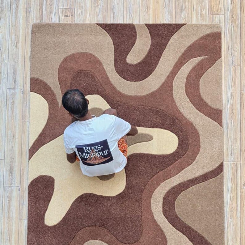 Op de afbeelding: Een rechthoekig vloerkleed met een wervelend patroon in bruin, beige en tan tinten. Het vloerkleed heeft een zacht, getextureerd uiterlijk. De persoon op de foto draagt een wit t-shirt met de tekst "Rugs Mirzapur".